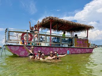 Group of friends enjoying a sunny beach day in shallow green water next to a pink floral pontoon tiki boat with a thatched roof, holding drinks and waving under a bright blue sky
