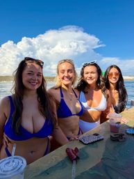 Four sun‑soaked women in bikinis smiling at a waterfront boat bar with phones and cold drinks on the counter, bright blue sky and large puffy clouds over the ocean — summer beach vibe.