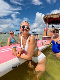 Smiling woman in white bikini with heart-shaped sunglasses leaning on a pink paddleboard in shallow coastal water, friends in swimsuits nearby, tiki-style party boat and blue sky — summer boat beach party