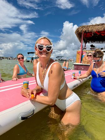 Smiling woman in white bikini with heart-shaped sunglasses leaning on a pink paddleboard in shallow coastal water, friends in swimsuits nearby, tiki-style party boat and blue sky — summer boat beach party