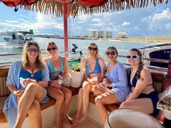 Five friends in colorful swimsuits and sunglasses relaxing under a tiki-style canopy on a leisure boat in a sunny coastal bay, with other boats, sandy shoreline and waterfront condos in the background.