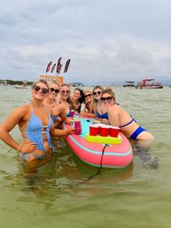Group of friends in bikinis smiling around a pink inflatable paddleboard with red solo cups and drinks in shallow coastal water, with anchored boats and beach flags in the background on an overcast summer day