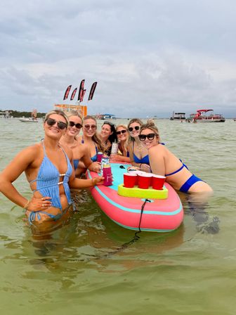 Group of friends in bikinis smiling around a pink inflatable paddleboard with red solo cups and drinks in shallow coastal water, with anchored boats and beach flags in the background on an overcast summer day