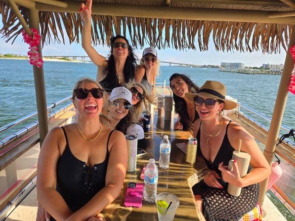 Seven women laughing and posing under a thatched tiki roof on a sunny boat cruise, drinks and water bottles on a wooden table, with a bridge and coastal buildings across the bay.