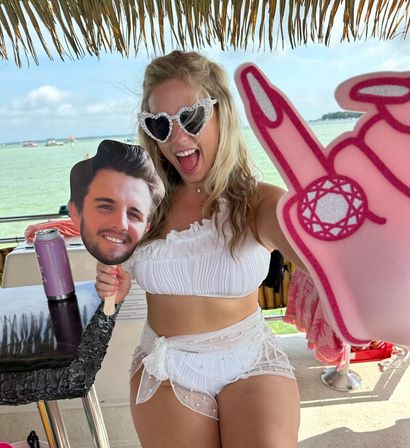Smiling woman in heart-shaped sunglasses and white bikini on a tiki-roof boat over green tropical water, holding a male face cutout and a pink foam hand with a ring