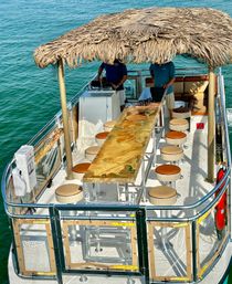 Tiki-roofed pontoon boat bar with glossy resin wood tabletop, round stools and cushioned lounge seating cruising turquoise coastal waters on a sunny day.