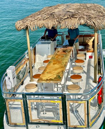 Tiki-roofed pontoon boat bar with glossy resin wood tabletop, round stools and cushioned lounge seating cruising turquoise coastal waters on a sunny day.