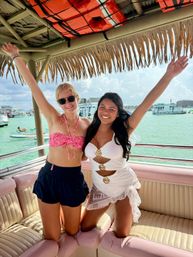 Two friends in swimwear cheer with arms raised under a thatched tiki canopy on a pink-cushioned boat, turquoise water and anchored boats in the background — tropical vacation boat party.