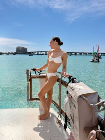 Smiling woman in a white bikini standing on a boat railing, gazing over clear turquoise tropical water toward a distant bridge, sailboats and resort buildings under a bright blue sky.