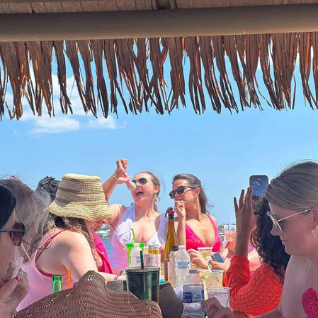Group of women at a sunny beachside bar under a thatched palapa, laughing and raising cocktails while one records on a smartphone against a clear blue sky and ocean backdrop.
