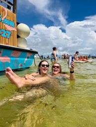 Two friends in sunglasses laughing and floating in shallow green beach water near a colorful flamingo-decorated boat, one holding a canned drink with paddleboarders and blue sky clouds in the background.