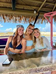 Three smiling friends in swimsuits leaning at a thatched-roof tiki beach bar with ocean water and sandy dunes under a bright blue sky, phone on the counter