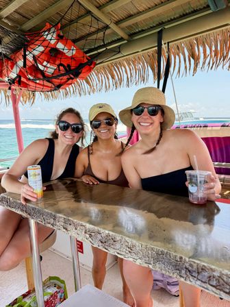 Three smiling friends in swimsuits and sunglasses at a tiki-style boat bar with a thatched roof, life jackets overhead, drinks on a weathered counter and turquoise ocean in the background — tropical boat vacation scene.