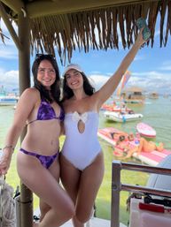 Two smiling women in swimsuits pose under a thatched tiki roof on a sunny tropical beach dock — one in a purple bikini and the other in a white one-piece holding a drink can, with paddleboards, boats and shallow turquoise water in the background.