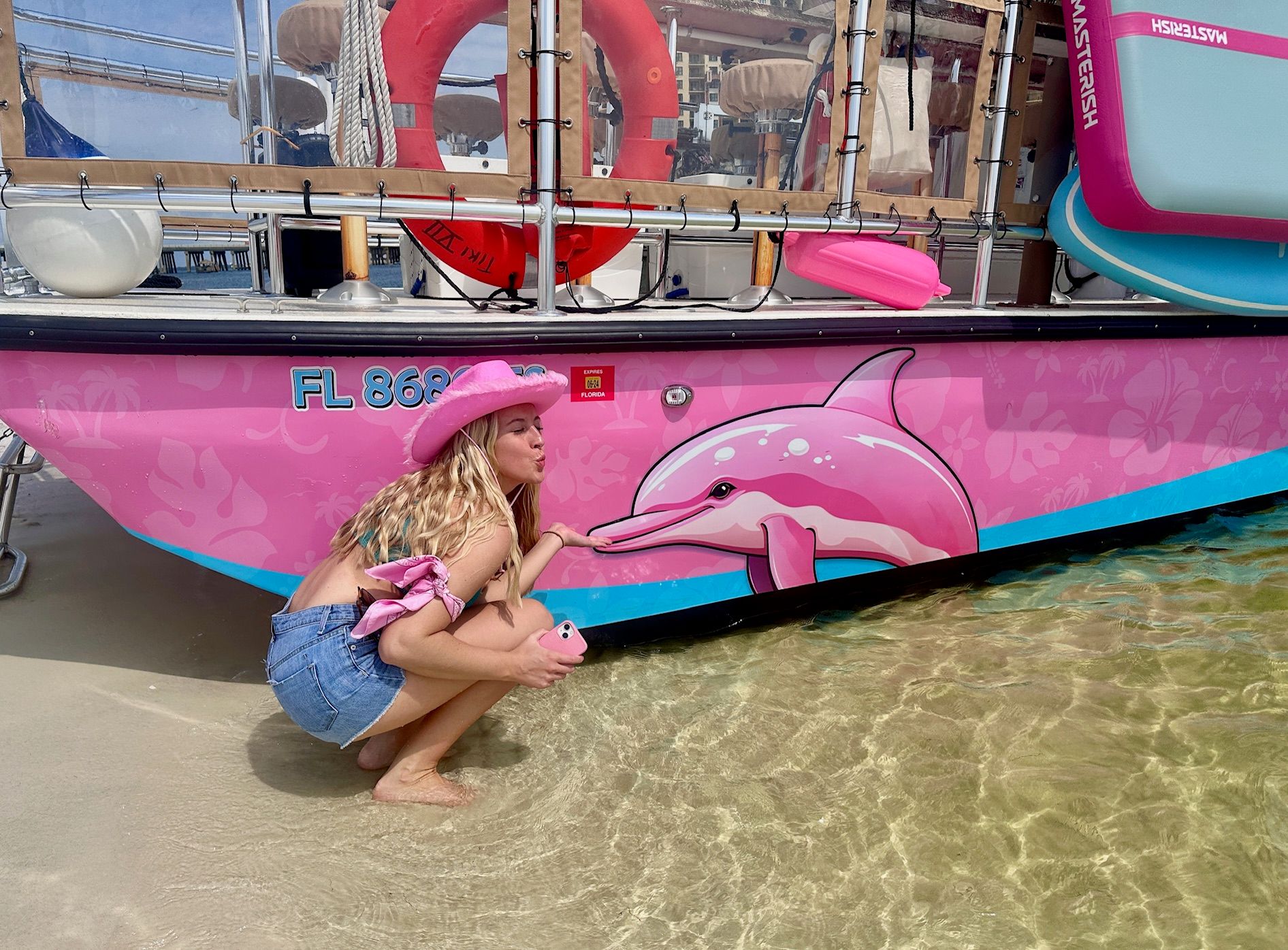 Person in a pink cowboy hat and denim shorts crouching in clear shallow water to 'kiss' a painted pink dolphin on the hull of a bright pink boat at a Florida beach