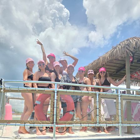 Group of women in black swimsuits and matching pink caps posing on a tropical boat deck by a thatched tiki roof, life rings visible and cloudy blue sky overhead.