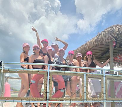 Group of women in black swimsuits and matching pink caps posing on a tropical boat deck by a thatched tiki roof, life rings visible and cloudy blue sky overhead.
