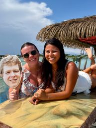 Two smiling women lounging at a tropical beach bar under a thatched palapa, one holding a face cutout with turquoise water and blue sky behind them.