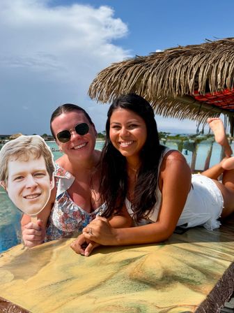 Two smiling women lounging at a tropical beach bar under a thatched palapa, one holding a face cutout with turquoise water and blue sky behind them.