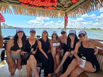 Seven friends in matching caps and black swimwear smiling on a tiki-roof pontoon boat, holding drinks with a sunny waterfront bay and marina in the background