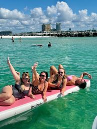 Four friends in swimsuits lounging and waving on a pink floating mat in clear turquoise beach water with a rocky shoreline and beachfront towers under a bright blue sky of puffy clouds, vacation scene.