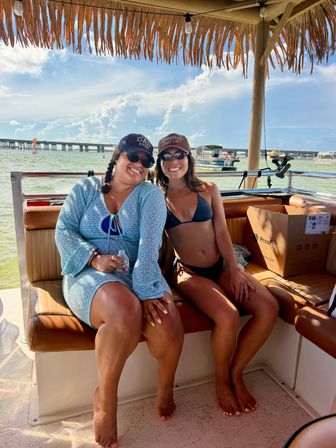 Two friends smiling in swimwear on a shaded pontoon boat under a tiki roof, sunny coastal bay with a bridge and other boats in the background.