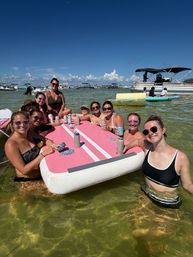 Group of friends enjoying a sunny sandbar beach day, smiling around a large pink inflatable floating table in shallow clear coastal water with anchored boats and blue sky in the background.
