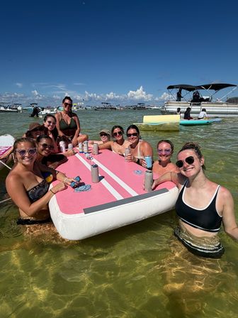 Group of friends enjoying a sunny sandbar beach day, smiling around a large pink inflatable floating table in shallow clear coastal water with anchored boats and blue sky in the background.