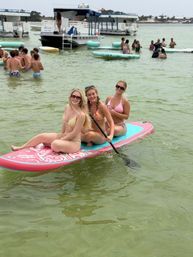 Three smiling women in bikinis seated on a pink-and-teal paddleboard holding a paddle in shallow green coastal water, with anchored party boats and groups of beachgoers on floating pads in the background