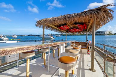 Tiki-style floating bar on a boat deck with thatched roof and round stools, overlooking a sunny marina with yachts on blue water.