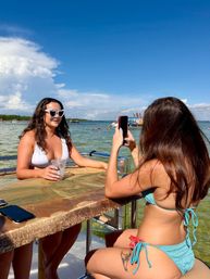 Two women in bikinis on a boat's waterfront bar — one posing in heart-shaped sunglasses with a drink while her friend snaps a photo against a bright blue sky and calm coastal bay.