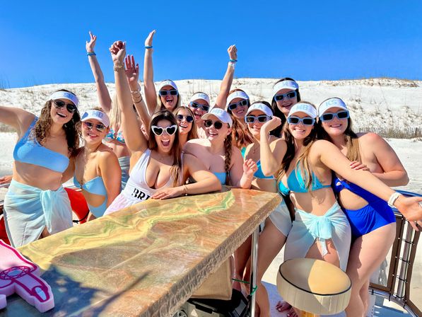 Energetic bachelorette party group of friends on a sunny white-sand beach, wearing swimsuits, visors and sunglasses with arms raised and a bride in a sash celebrating together