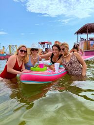 Four friends in swimsuits smiling around a bright pink inflatable float in shallow beach water, holding drinks with a tiki hut and water slides in the sunny blue-sky background — summer beach float party.