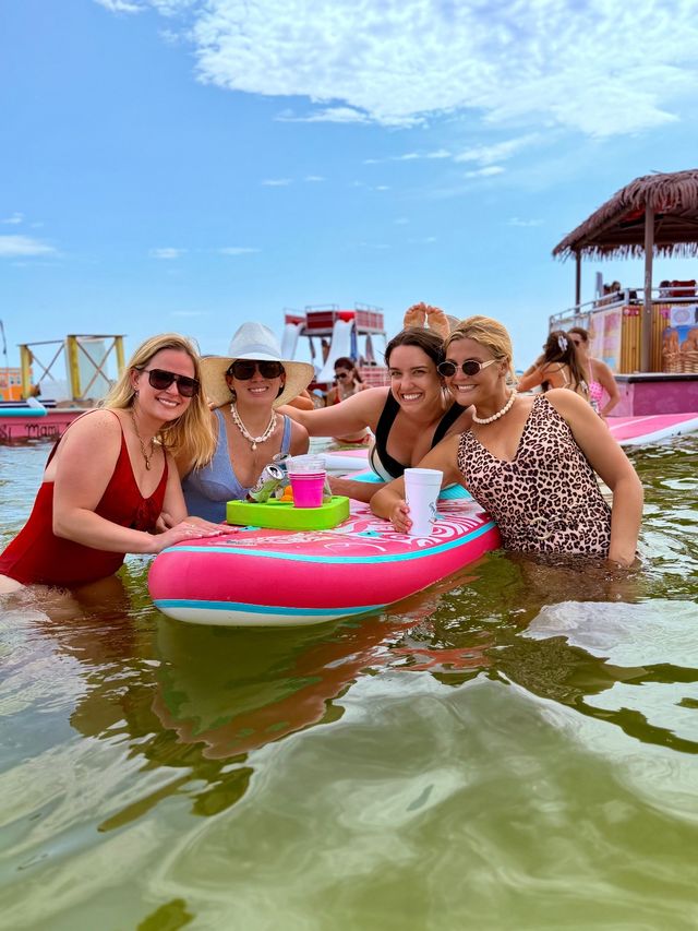 Four friends in swimsuits smiling around a bright pink inflatable float in shallow beach water, holding drinks with a tiki hut and water slides in the sunny blue-sky background — summer beach float party.