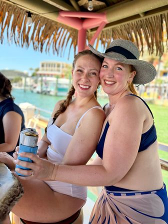 Two smiling friends in swimwear at a dockside tiki bar by a marina — one wearing a sun hat, the other holding a canned drink in a blue koozie with turquoise water and boats in the background.