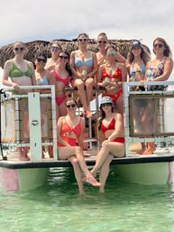 Group of women in colorful swimsuits and sunglasses posing and smiling on a pontoon boat with a tiki-style canopy above clear turquoise water, some holding canned drinks on a sunny summer day.