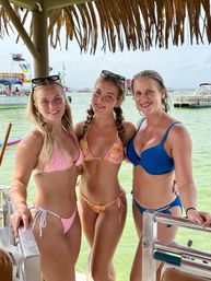 Three smiling women in colorful bikinis standing under a thatched tiki roof on a boat with clear green water, nearby boats and beach activity in the background — fun tropical vacation vibe.