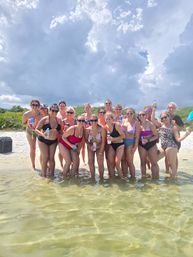 Group of women in colorful swimsuits standing waist-deep in clear shallow water on a sandy beach, holding drinks and smiling with green dunes behind them and dramatic summer clouds overhead.
