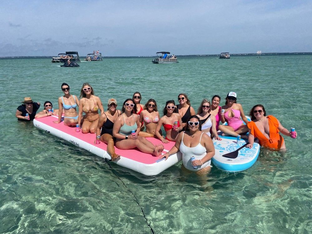Group of friends in colorful swimsuits lounging on a large pink inflatable and a blue paddleboard over clear turquoise sandbar water, holding drinks with boats anchored on the horizon on a sunny day.