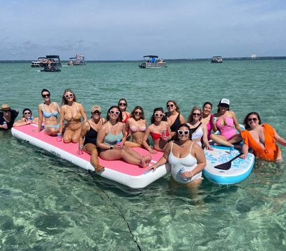 Group of friends in colorful swimsuits lounging on a large pink inflatable and a blue paddleboard over clear turquoise sandbar water, holding drinks with boats anchored on the horizon on a sunny day.