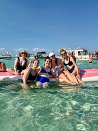 Smiling group of five friends in swimsuits lounging on a pink floating paddleboard in clear turquoise shallow water with party boats and blue sky on a sunny beach day