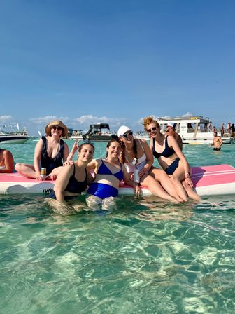 Smiling group of five friends in swimsuits lounging on a pink floating paddleboard in clear turquoise shallow water with party boats and blue sky on a sunny beach day
