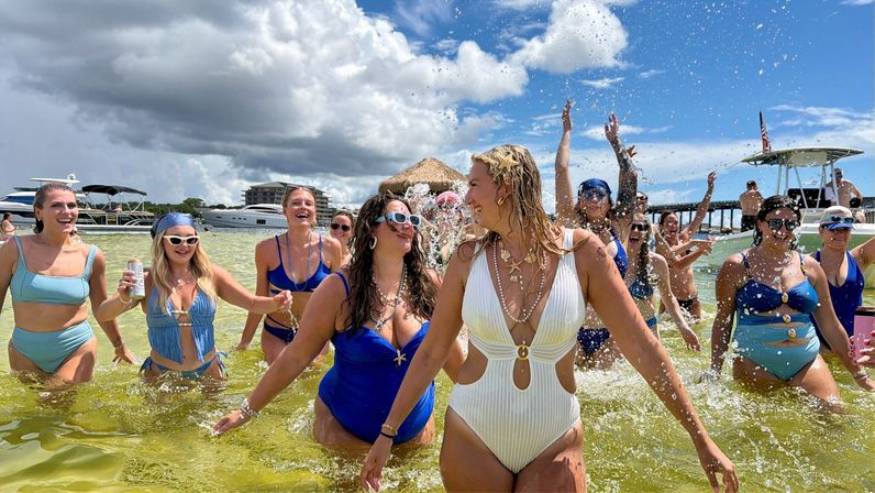 Group of women in colorful swimsuits laughing and splashing in shallow green coastal water at a sandbar with yachts, boats and a pier under a sunny, partly cloudy sky