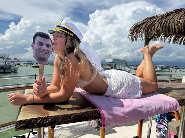 Bachelorette in a white "bride" captain hat and veil lounging on a dock table, kissing a face cutout on a stick with boats, a palapa umbrella, and dramatic cloudy sky over a busy bay