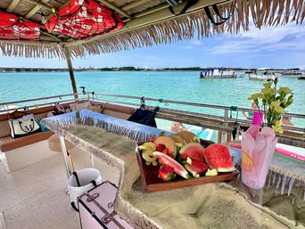 Sunny thatched-roof tiki boat deck overlooking a turquoise bay with anchored boats, a wooden tray of sliced watermelon and melon, pink utensils, and red life jackets stored overhead