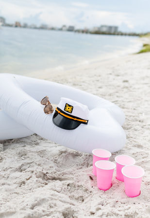 White inflatable tube on a sandy beach by the shoreline with a captain's hat and sunglasses on top and four pink party cups nearby — playful oceanfront beach party scene.