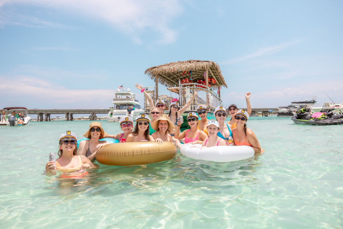 Smiling group of women in captain hats on inflatable tubes in clear turquoise water beside a tiki-roof party boat, with yachts, jet skis and a bridge under a sunny sky.