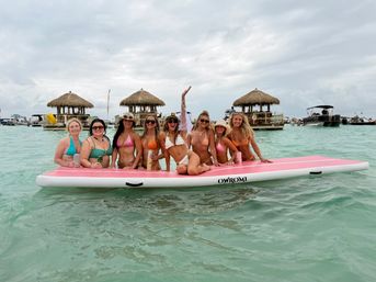 Eight women in colorful bikinis posing on a large pink inflatable float in shallow turquoise water near tiki-hut floating bars and anchored boats under a cloudy tropical sky