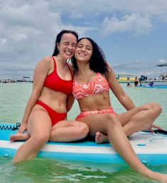 Two smiling women in red swimsuits sitting close on a paddleboard in shallow turquoise water, hugging and posing with boats and a partly cloudy sky in the background — sunny beach day vibe.