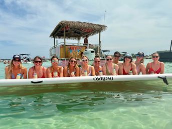 Group of smiling women on a long inflatable float in shallow turquoise water, holding canned drinks with a tiki‑roof party boat in the background on a sunny beach day.